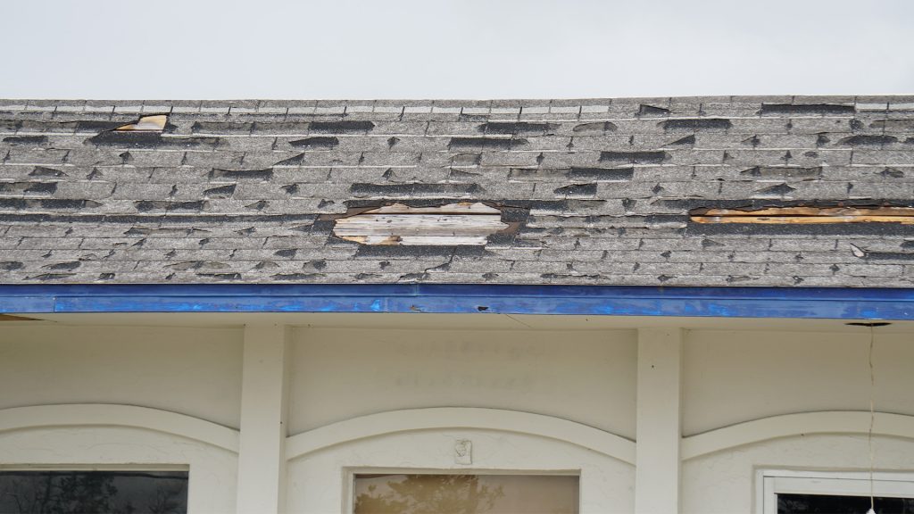 Dilapidated roof with warped asphalt shingles and a large torn area exposing underlayment, over a white building with blue trim.