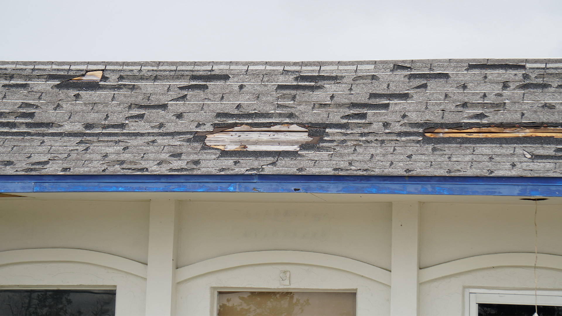 Hurricane Isaias Claim Lawyer - Gelber Law Group Dilapidated roof with warped asphalt shingles and a large torn area exposing underlayment, over a white building with blue trim.