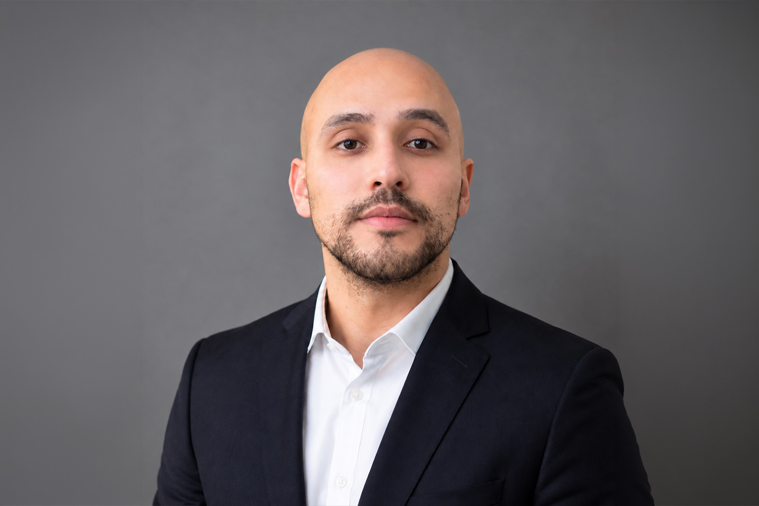 Portrait of a bald man in a dark blazer and white shirt against a gray backdrop, looking at the camera.